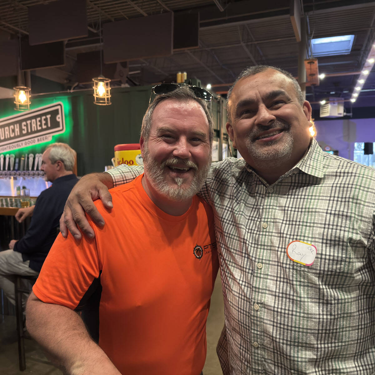 Two smiling men stand arm-in-arm in a bustling brewery, capturing the festive spirit of Oktoberfest 2024. One wears an orange shirt, the other a checked shirt with a name tag. In the background, patrons enjoy drinks under hanging lights at the bar, adding to the vibrant atmosphere.