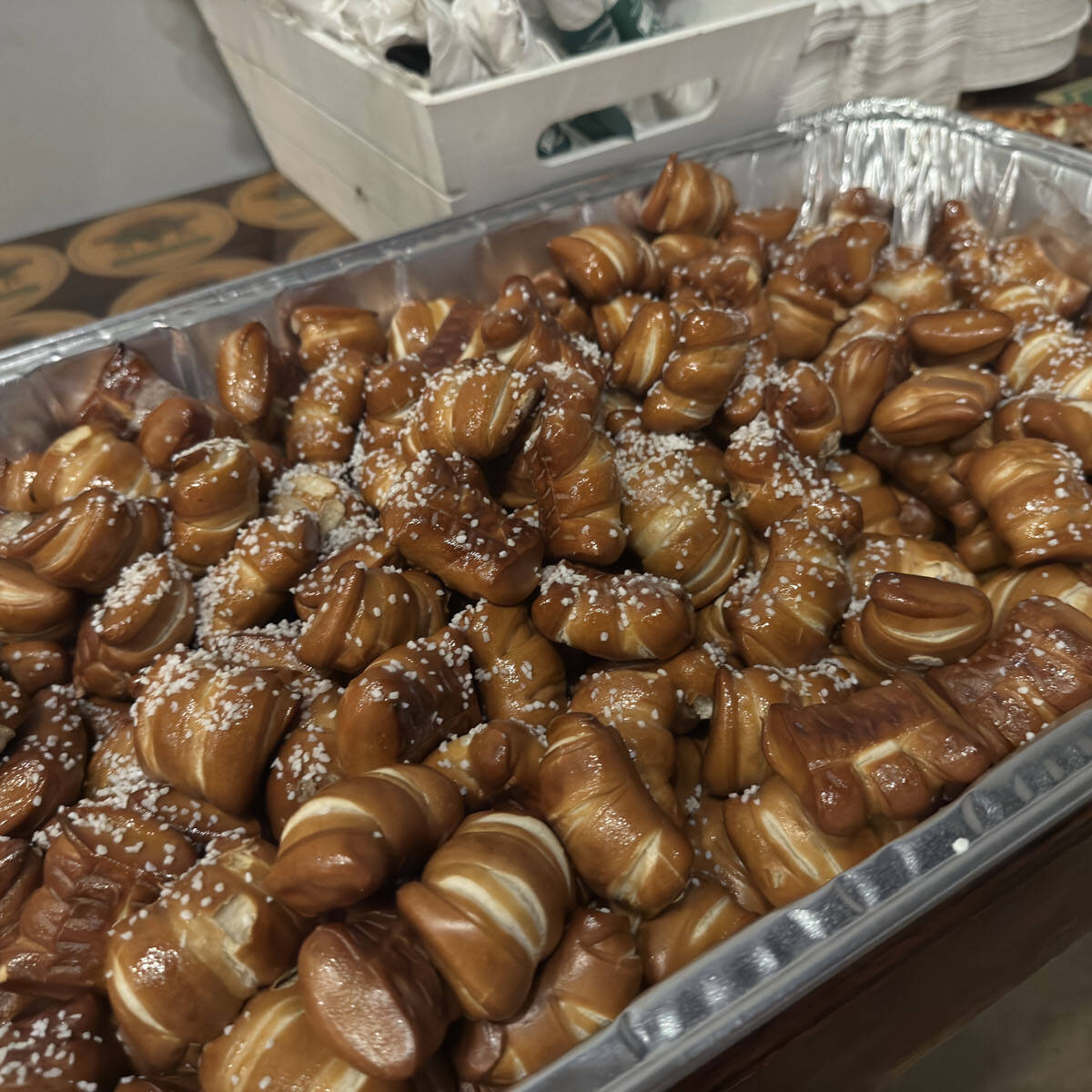 A large tray filled with mini pretzels, topped with coarse salt, perfect for Oktoberfest 2024. The pretzels are golden brown and clustered together. In the background, there is a glimpse of another dish and a white container.