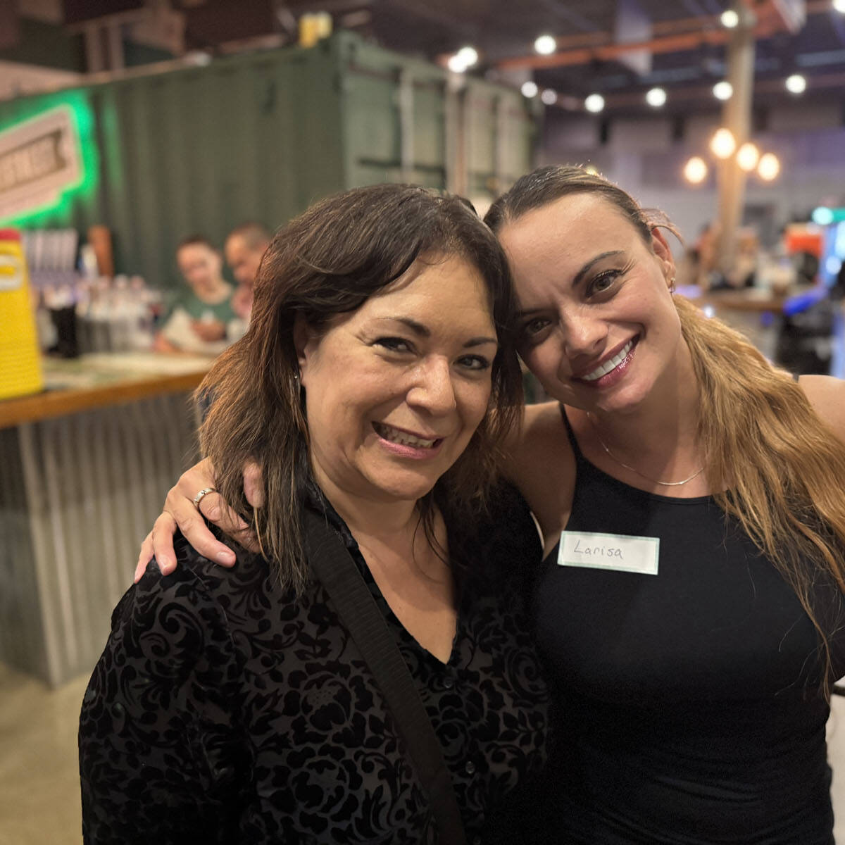 Two women smiling and posing for a photo in a lively indoor setting reminiscent of Oktoberfest, with a bar and cheerful people in the background. One woman, sporting long hair and a name tag, captures the festive 2024 spirit. The atmosphere is casual and social.