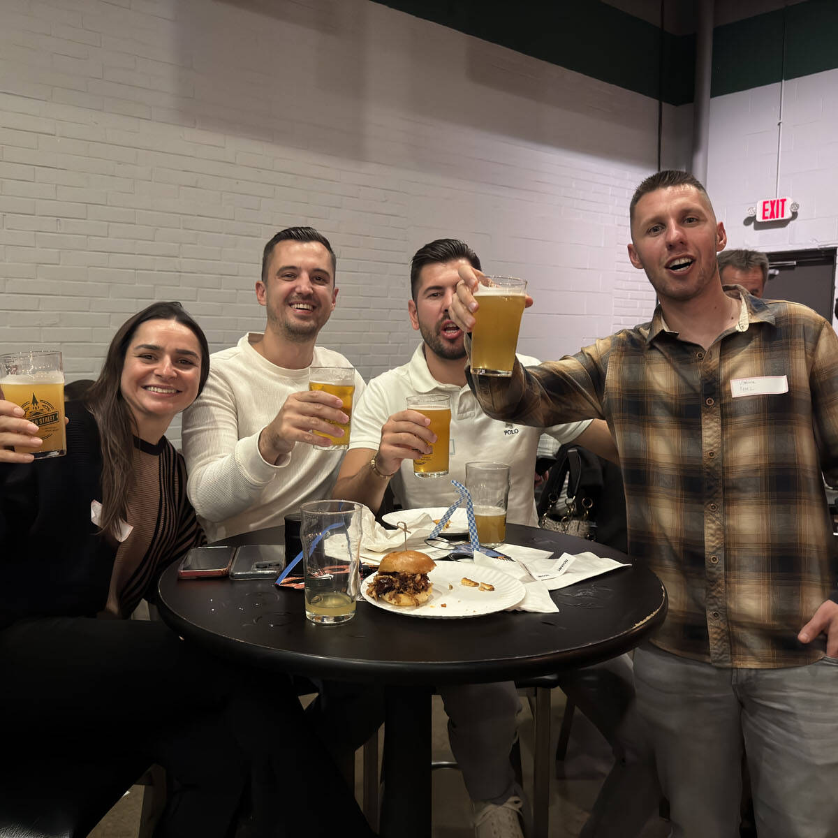 Four people sitting at a round table in a lively bar, raising their drinks and smiling at the camera, capturing the spirit of Oktoberfest 2024. The table is adorned with snacks, a glass, and napkins. Others in the background are also reveling in the festive atmosphere.