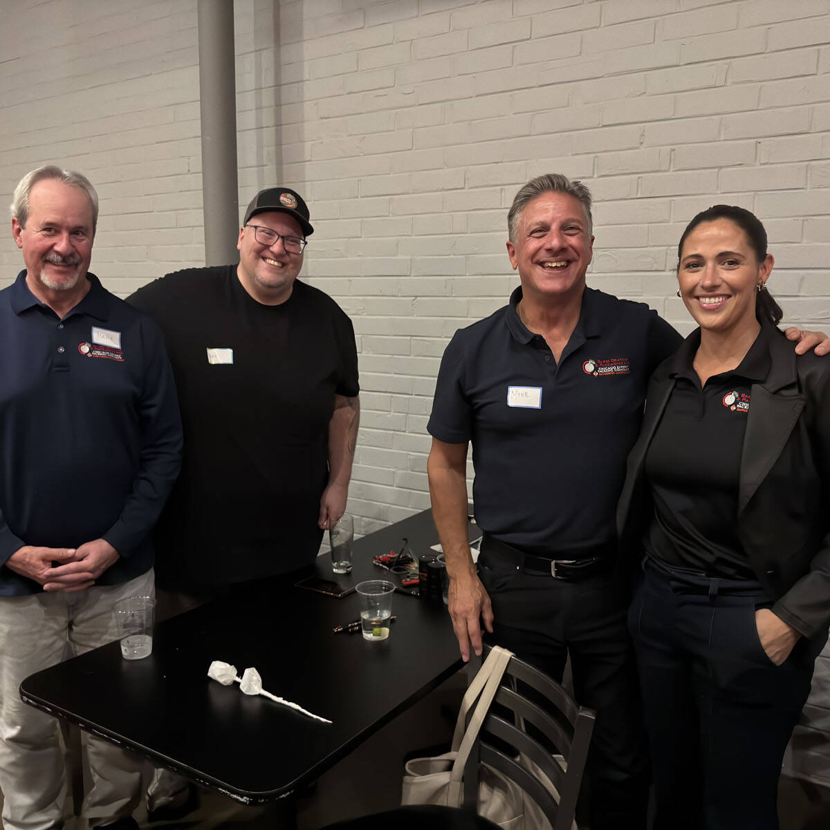 Four people smiling and standing together in a casual setting with a table in front of them, dressed in dark attire and sporting name tags. The light brick wall hints at an Oktoberfest 2024 event vibe.