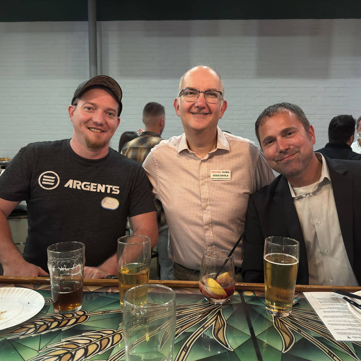 Three men are sitting together at a bar, smiling amidst the lively atmosphere of Oktoberfest 2024. They have drinks in front of them, including beer and cocktails. The middle man is wearing glasses and a name tag. A few other people are visible in the background, but out of focus.