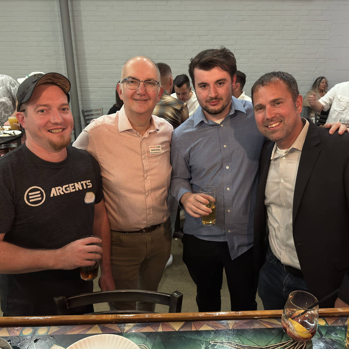 Four men smiling at a social gathering, holding drinks. They are standing close together in a brightly lit room, capturing the spirit of Oktoberfest. In the background, others are mingling while the table in front is adorned with empty plates and glasses.