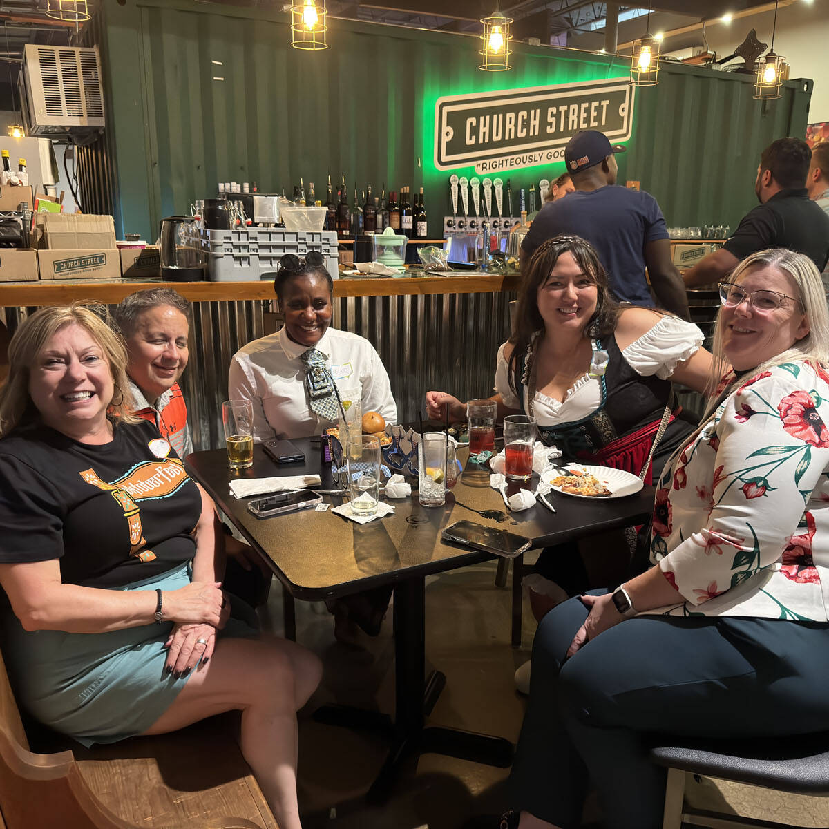 Five people are seated around a table at a bar, smiling at the camera. The background reveals a bar counter with beverage taps and a sign reading "Church Street," capturing the early excitement for Oktoberfest 2024 events.