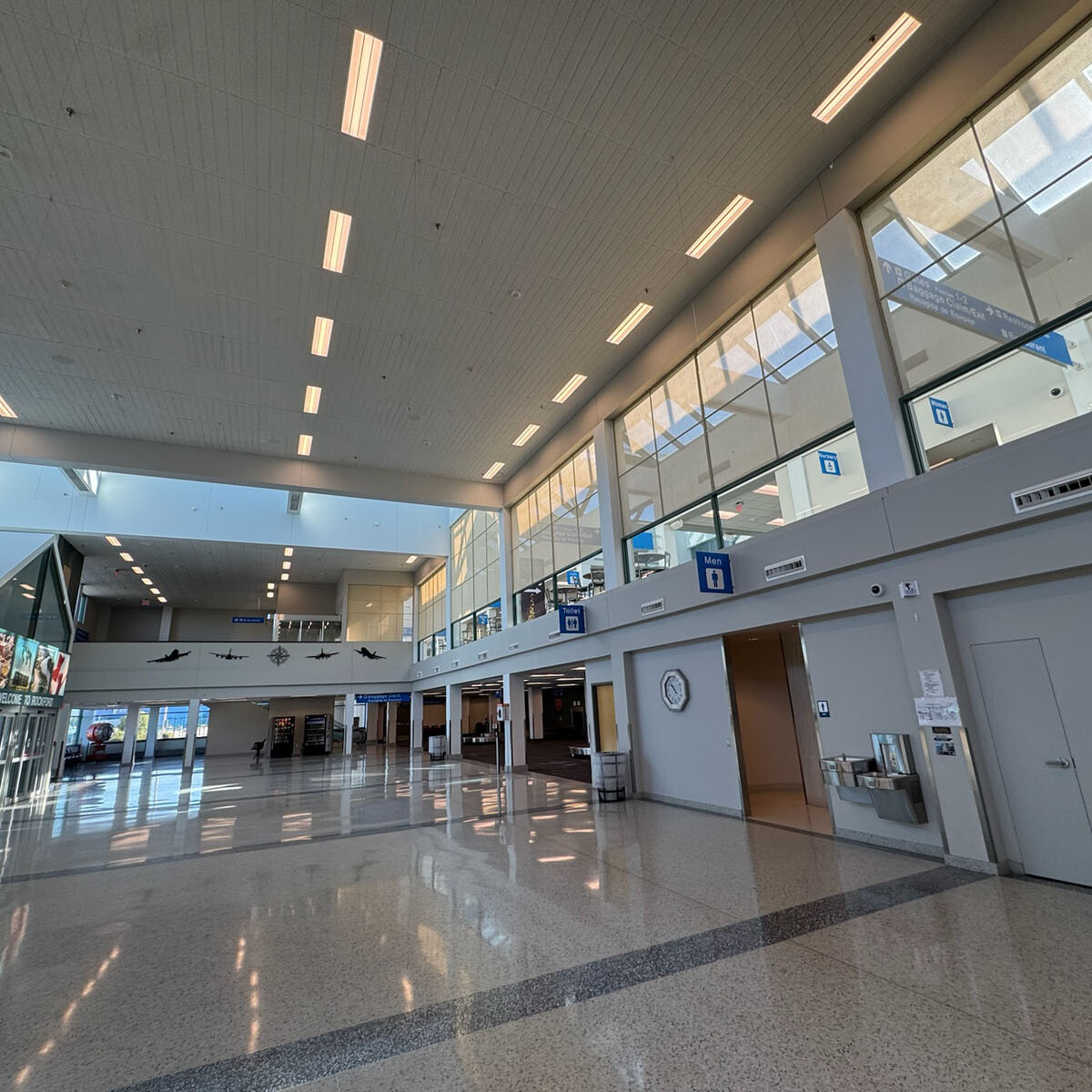 A spacious, modern airport terminal with high ceilings and large windows welcomes travelers to Rockport. Bright overhead lighting illuminates the clean, polished floors as signs guide eager visitors, while small shop entrances offer enticing glimpses into local delights.