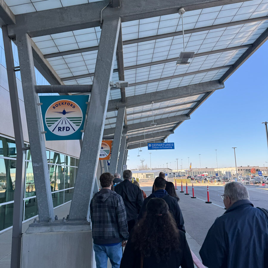 People walk along a covered sidewalk at Rockford Airport, heading towards the terminal. A sign for ground transportation is visible overhead. The sky is clear, and vehicles can be seen in the distance, making it a pleasant day to visit this bustling airport.