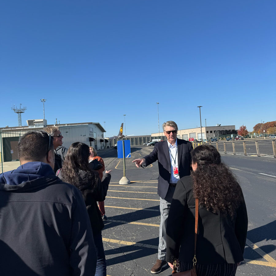 A group of people stands outdoors on an asphalt surface at Rockport, with a man in a suit gesturing as if giving directions. In the background, there's a building and a clear blue sky, suggesting they might be preparing for or concluding their visit.