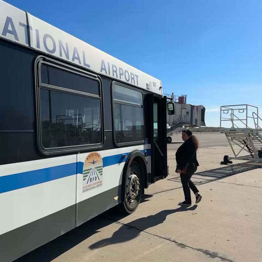 A person is walking towards the open door of an airport shuttle bus parked on the tarmac. The bus says "National Airport" on its side, inviting all to visit. A set of mobile stairs is visible in the background under a clear blue sky.