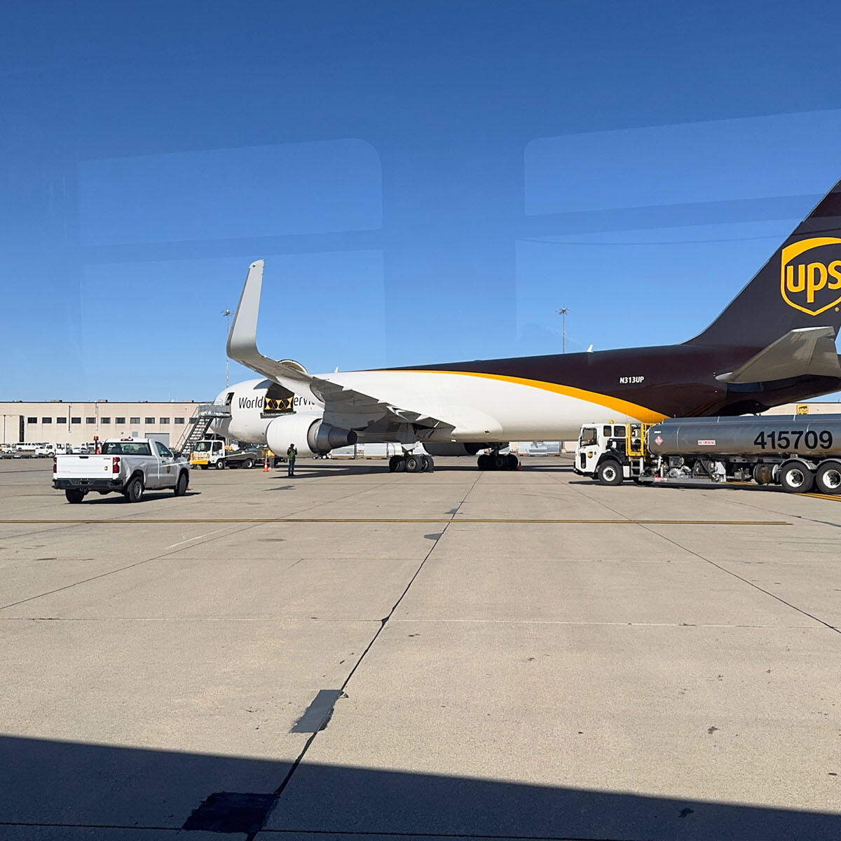 A UPS cargo plane is parked on an airport tarmac at Rockport Airport under a clear blue sky. A fuel truck is positioned near the plane's wing, and a small service vehicle is nearby. The airport terminal, perfect for those planning to visit, is visible in the background.