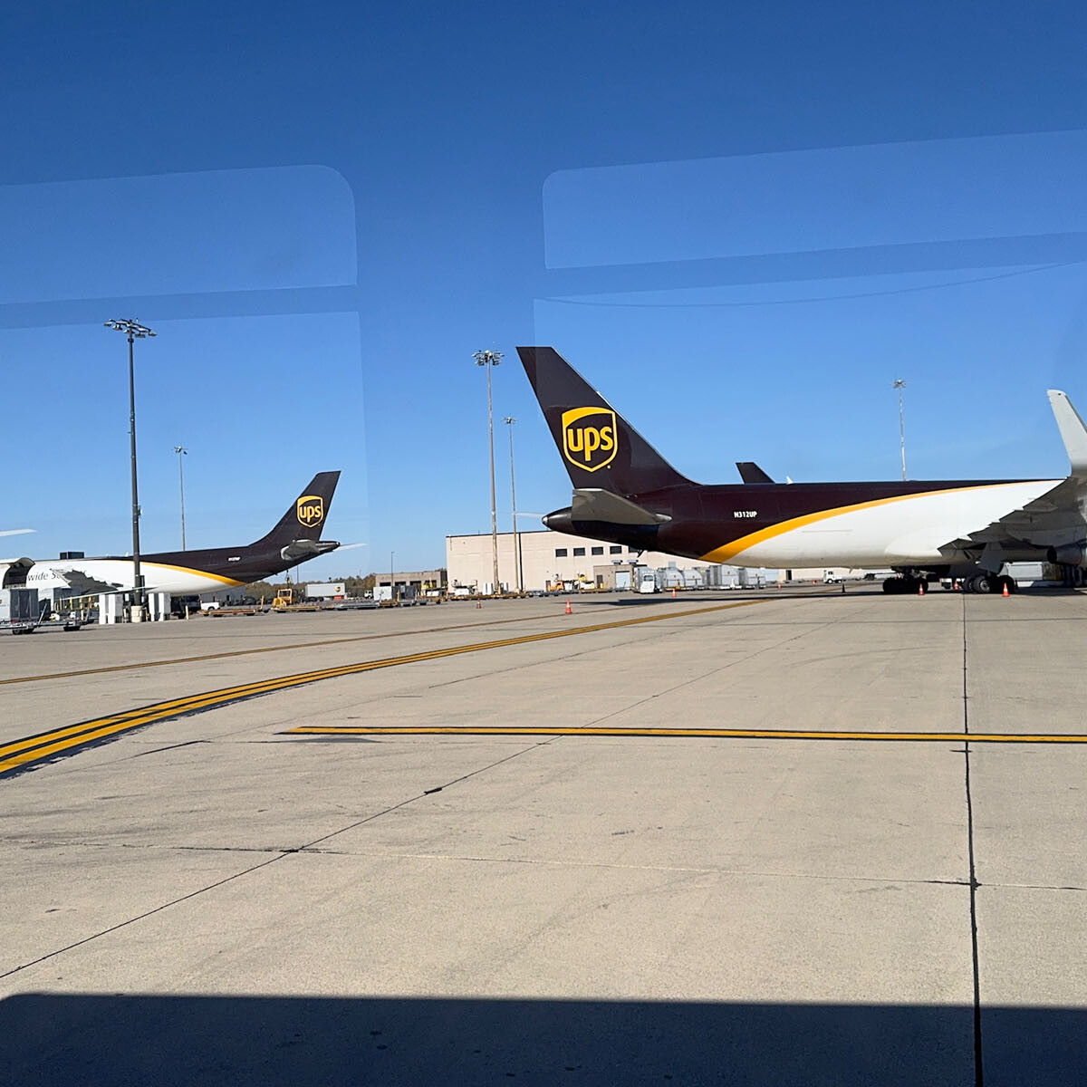 UPS cargo planes parked on a bustling Rockport airport tarmac under a clear blue sky. The planes, showcasing the iconic UPS logo with their distinct white and brown coloring, stand ready for action. In the background, several airport buildings and a clear runway invite a visit of awe for aviation enthusiasts.