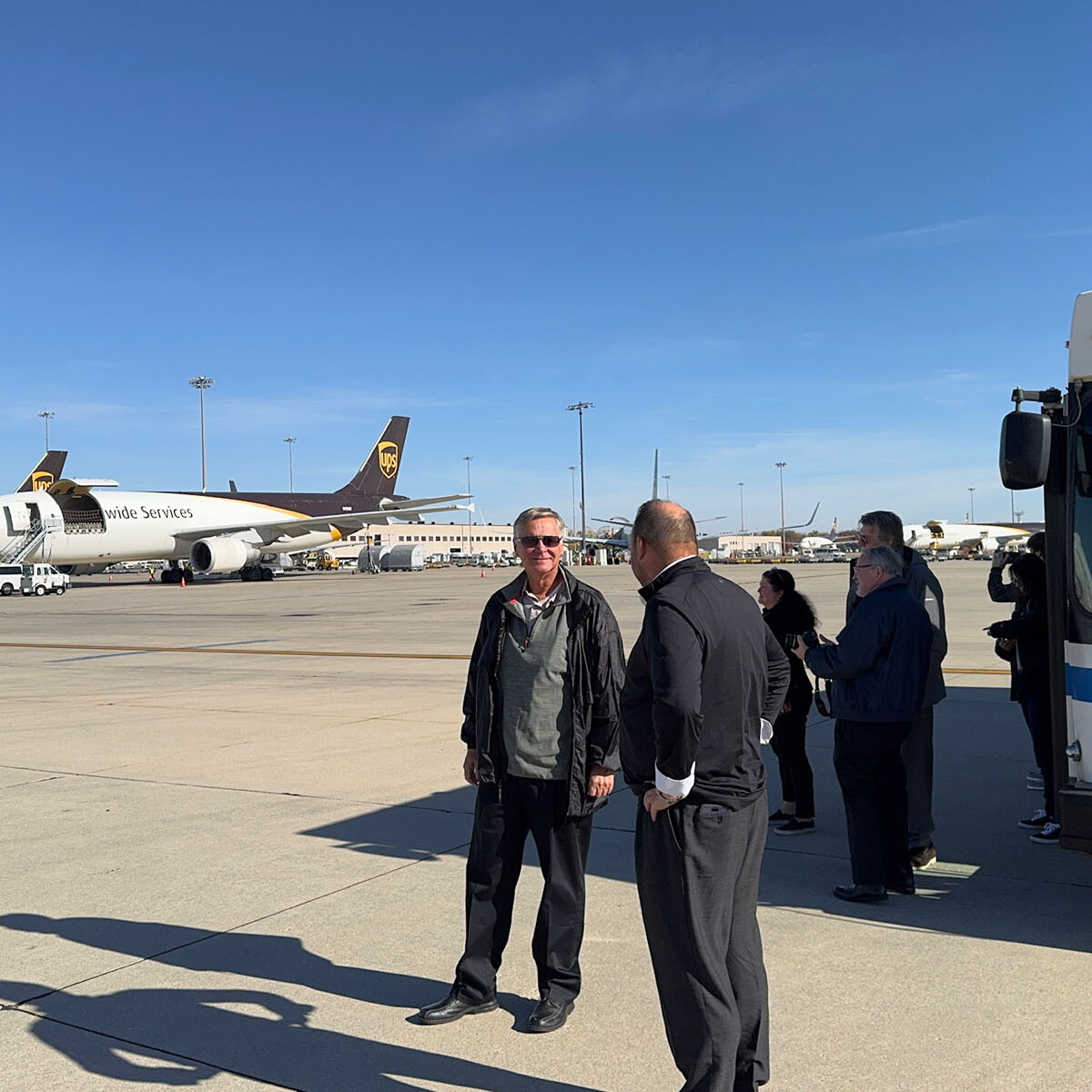 People standing on an airport tarmac near parked UPS planes and a shuttle bus. The clear blue sky sets the scene for transit activity. Some are engaged in conversation, perhaps discussing their visit to Rockport or planning their next destination in this bustling airport environment.