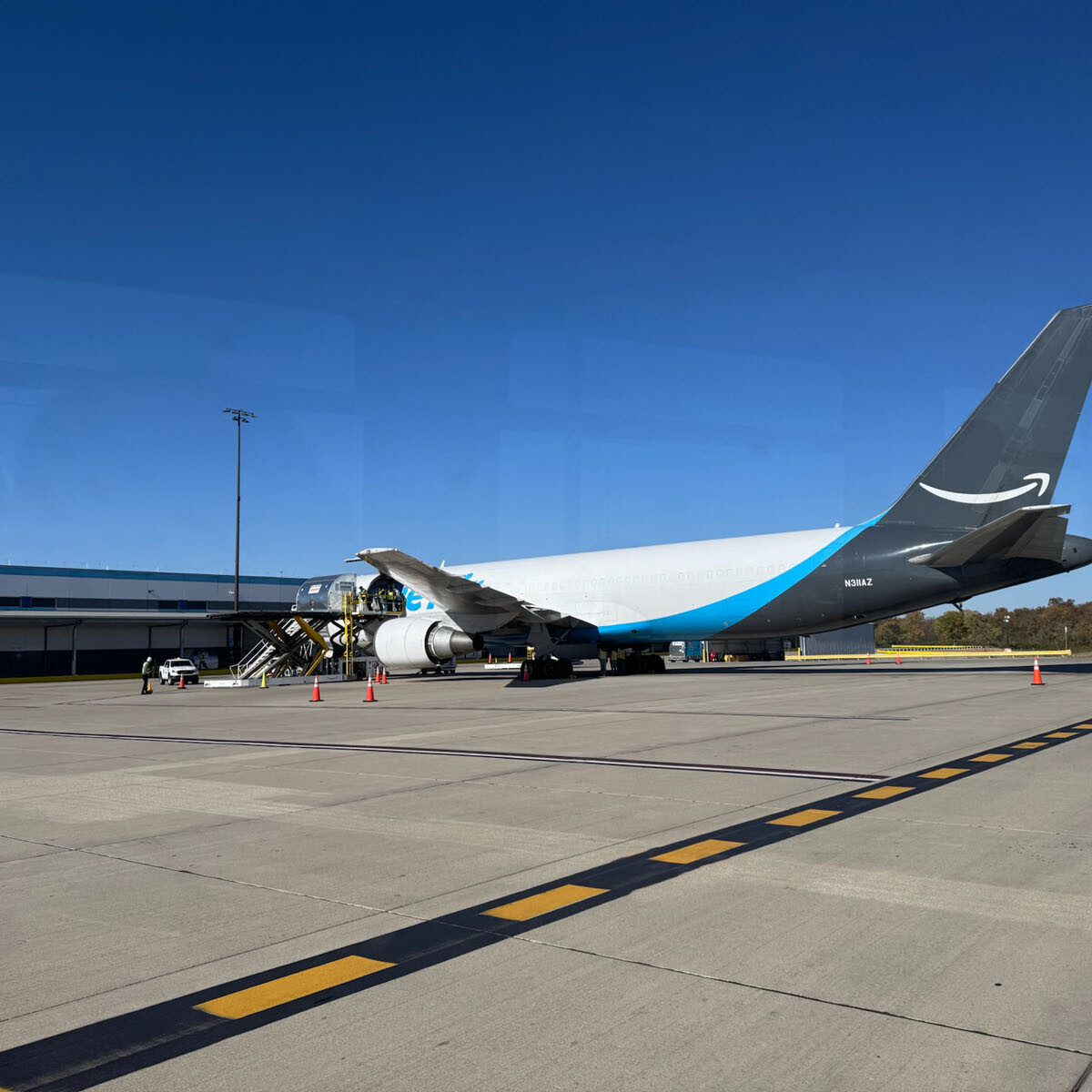 A cargo airplane with a blue and white tail is parked on the Rockport airport tarmac. Stairs are positioned by the aircraft's door, with several vehicles and cones scattered around. A clear blue sky forms the perfect backdrop for visitors.