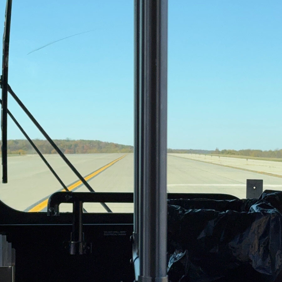 View from inside a bus on an open road under a clear blue sky, the perfect start to a Rockport visit. Framed by the bus window, the road ahead is lined with trees in the distance, promising adventure beyond just an airport layover.