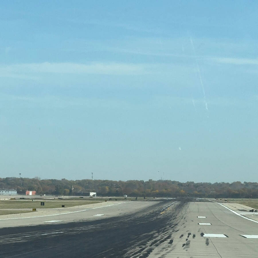 A concrete runway stretches into the distance at Rockport Airport, with tire marks visible on the surface. The sky is clear, and trees can be seen in the background under a bright blue sky. It's a perfect day to visit.