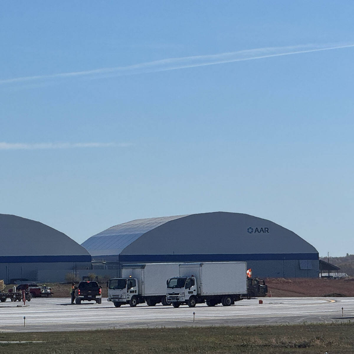 A large, open area at Rockport Airport showcases two hangars displaying "AAR." Nearby, several trucks and a marked car are parked with a portable toilet visible in the foreground. The sky invites you to visit under its clear expanse dotted with scattered clouds.