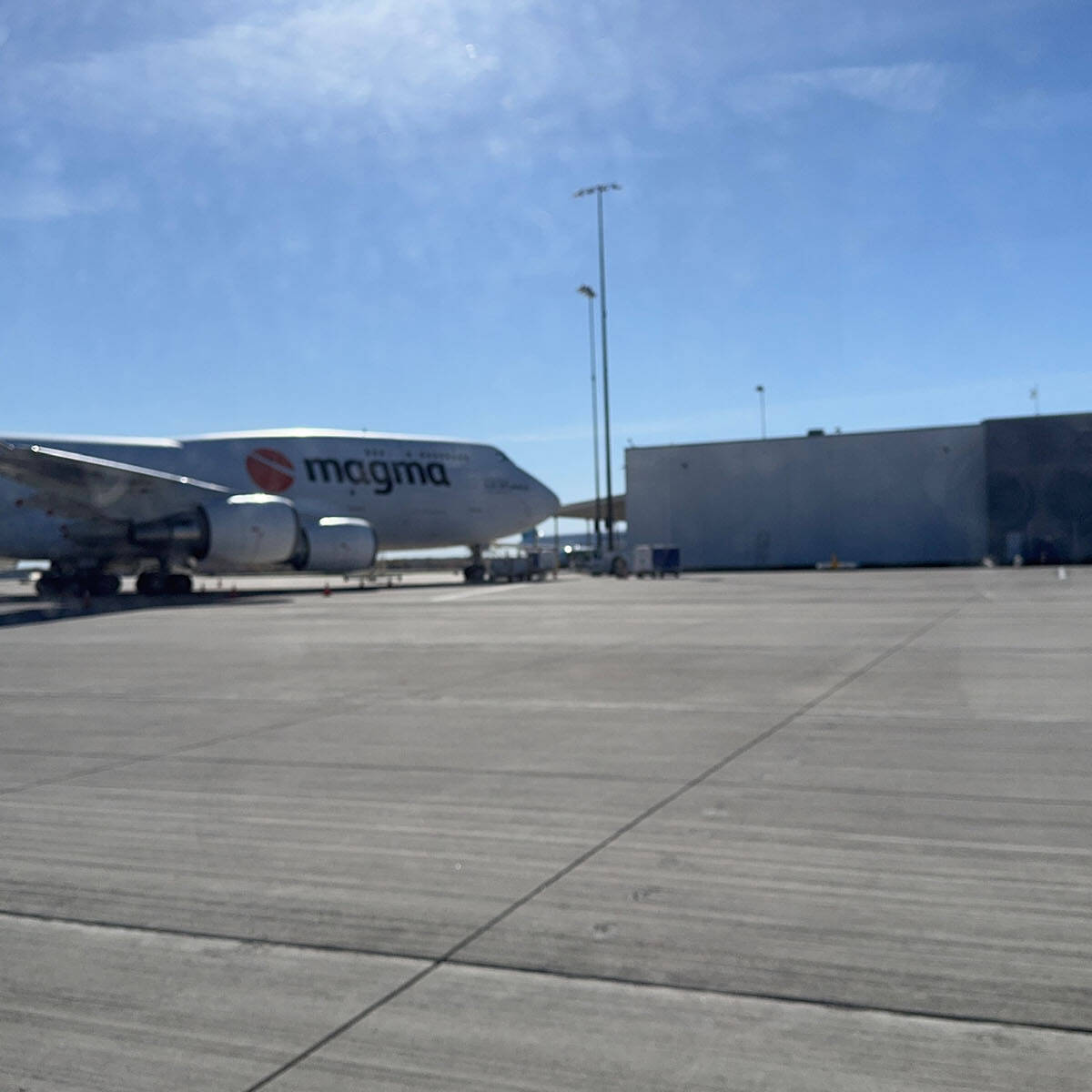 A cargo airplane with "magma" emblazoned on the fuselage is parked on the airport tarmac near a large gray building, set against a clear blue sky. It's an impressive sight for anyone who visits this bustling hub.