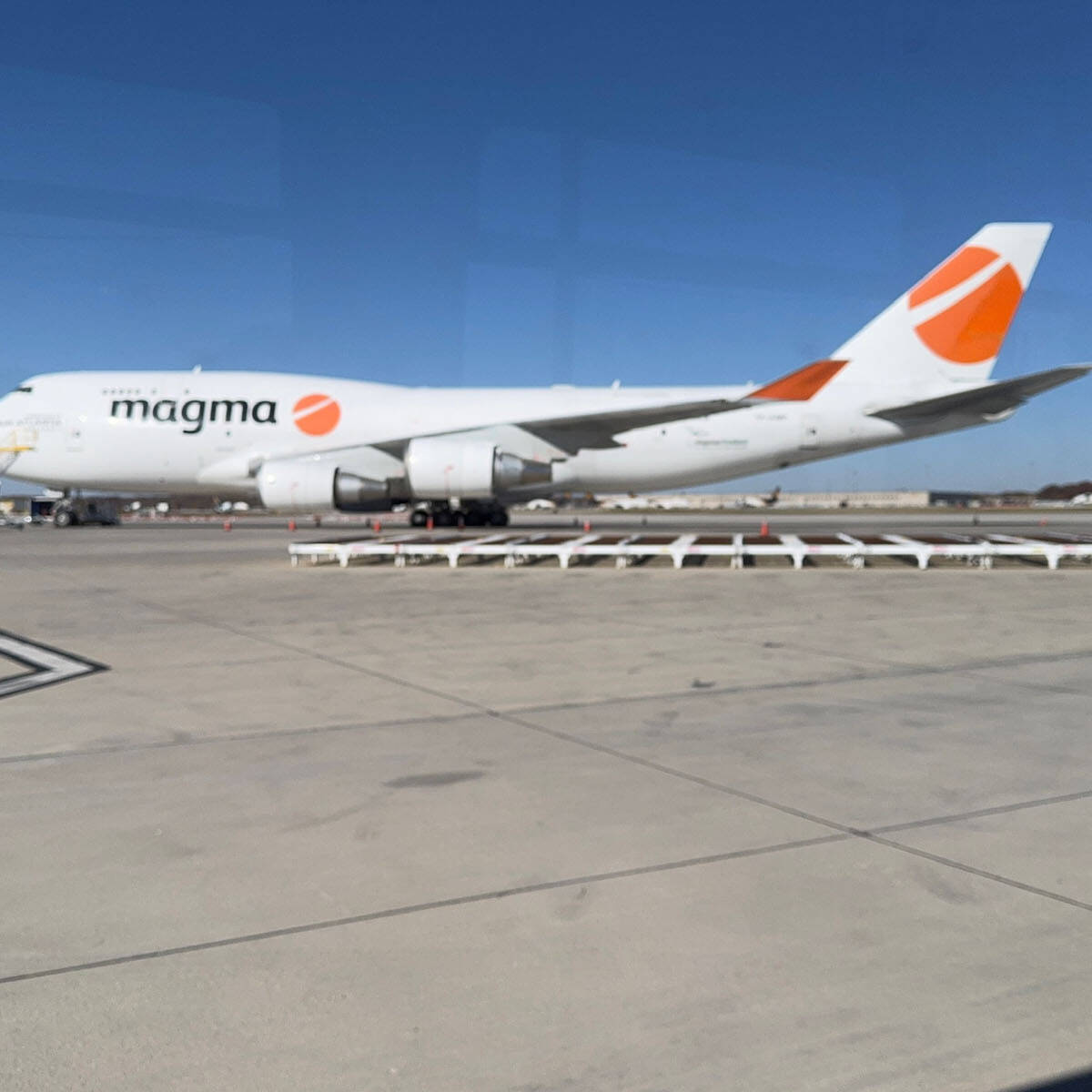 A large white cargo airplane with the logo "magma" and an orange design on its tail is parked on the tarmac at Rockport Airport on a clear day. The sky is blue, with no visible clouds, inviting visitors to explore the area.