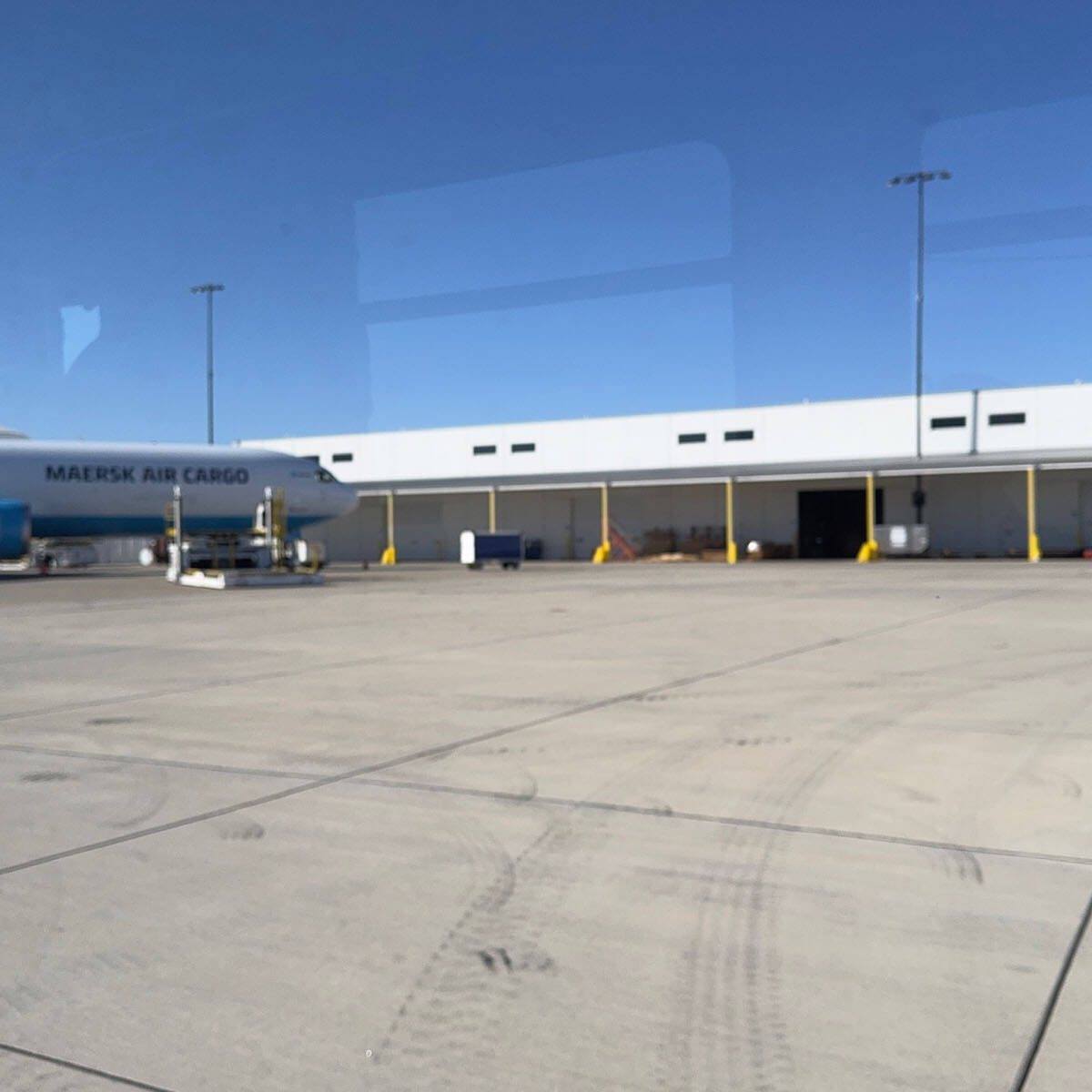 A cargo plane is parked on the expansive tarmac at Rockport Airport, in front of a large white warehouse with multiple bay doors. The sky is clear and blue, and there are yellow barriers near the building, welcoming visitors to this bustling hub.