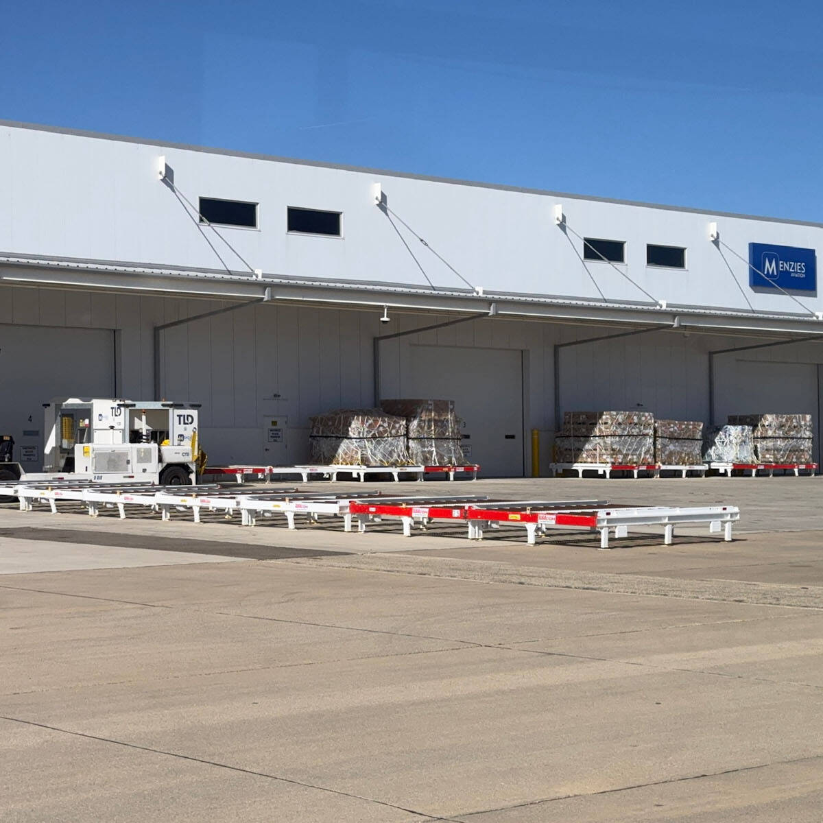 A large warehouse with a white exterior and a blue Rockport logo is visible. Several stacks of crates or materials are lined up along the building. A small industrial vehicle and a forklift are parked in the foreground on a concrete surface, making it ideal for those who visit frequently.