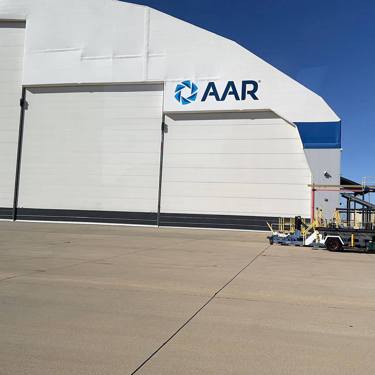 A large industrial building with the logo "AAR" greets visitors in Rockport on a clear day. The foreground highlights a wide concrete surface, reminiscent of an airport runway, with various vehicles and equipment parked nearby.