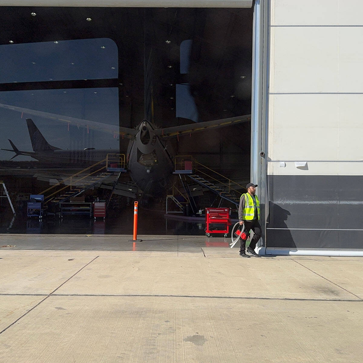 At Rockport Airport, an airplane sits inside a hangar undergoing maintenance. A worker in a yellow safety vest walks along the exterior of the hangar. Various equipment and tools are visible near the airplane under bright, clear skies—a scene inviting any aviation enthusiast to visit.