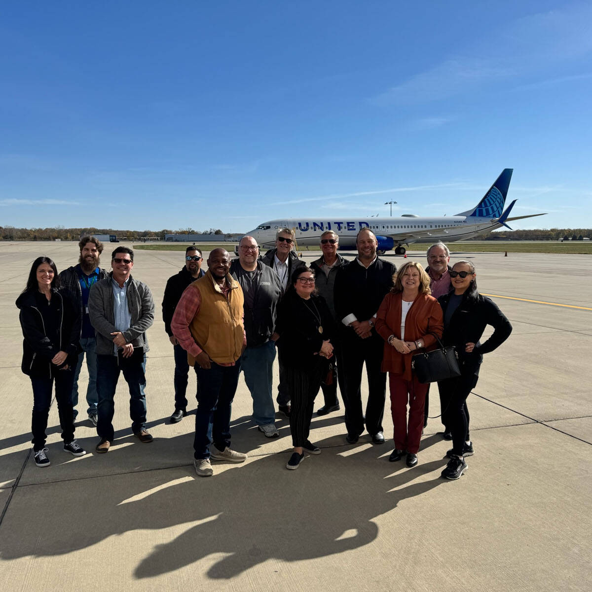 A group of people stands on an airport tarmac with a United airplane in the background, ready to visit new destinations. The sky is clear and blue as they casually pose for a photo, capturing the excitement of their impending journey.