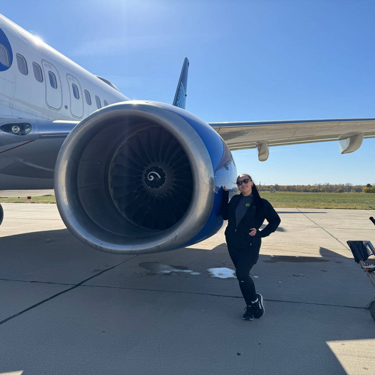 A person in dark clothing and sunglasses strikes a pose with one hand on their hip in front of a large airplane engine at the bustling Rockport airport. The sky is clear and sunny, with a small equipment cart adding to the vibrant scene.