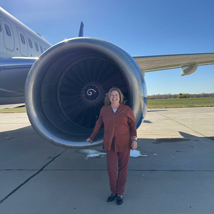 A person in a reddish-brown outfit stands beside the engine of an airplane at the airport on a sunny day, with a clear blue sky and grassy horizon in the background, capturing the spirit of adventure before embarking on their visit to Rockport.