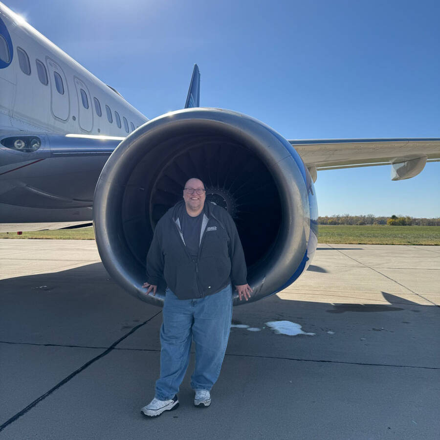 A person stands inside the opening of a large airplane engine, smiling on a sunny day at the airport. The aircraft is parked on the tarmac, and the sky is clear and blue, making it a perfect time to plan your next visit.