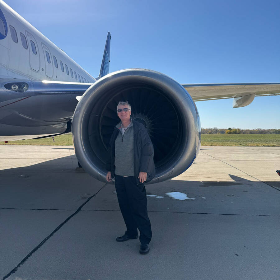 A person stands on a sunny day at the airport next to the engine of a large airplane on a tarmac. The sky is clear and blue. The person, perhaps visiting Rockport or another destination, is wearing sunglasses, a jacket, and dark pants.