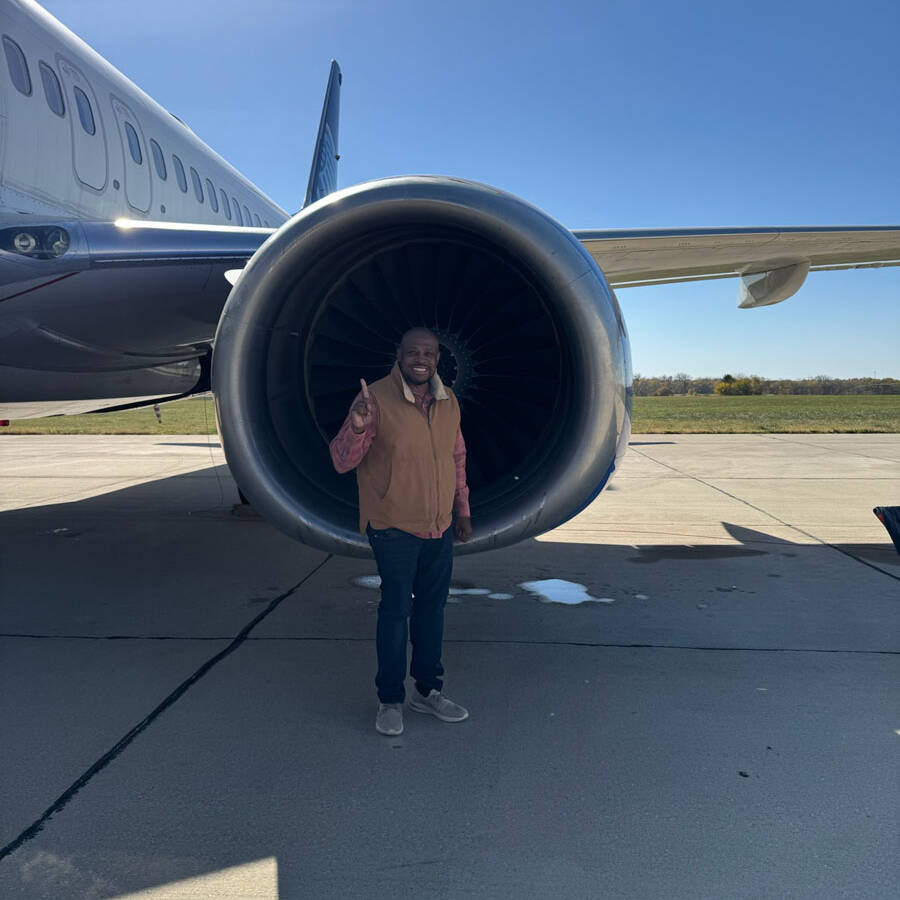 A person stands smiling next to a large airplane engine on a sunny day at the airport. Wearing a tan vest and jeans, they capture the essence of their visit as the aircraft's wing and tail stretch out in the background.