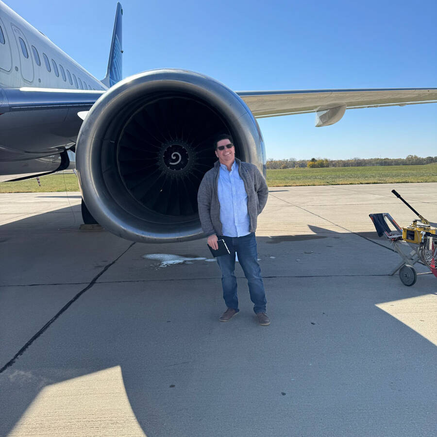 A person wearing sunglasses and a jacket stands on the tarmac at Rockport Airport, in front of a large airplane engine, with the aircraft’s wing stretching under a clear blue sky.