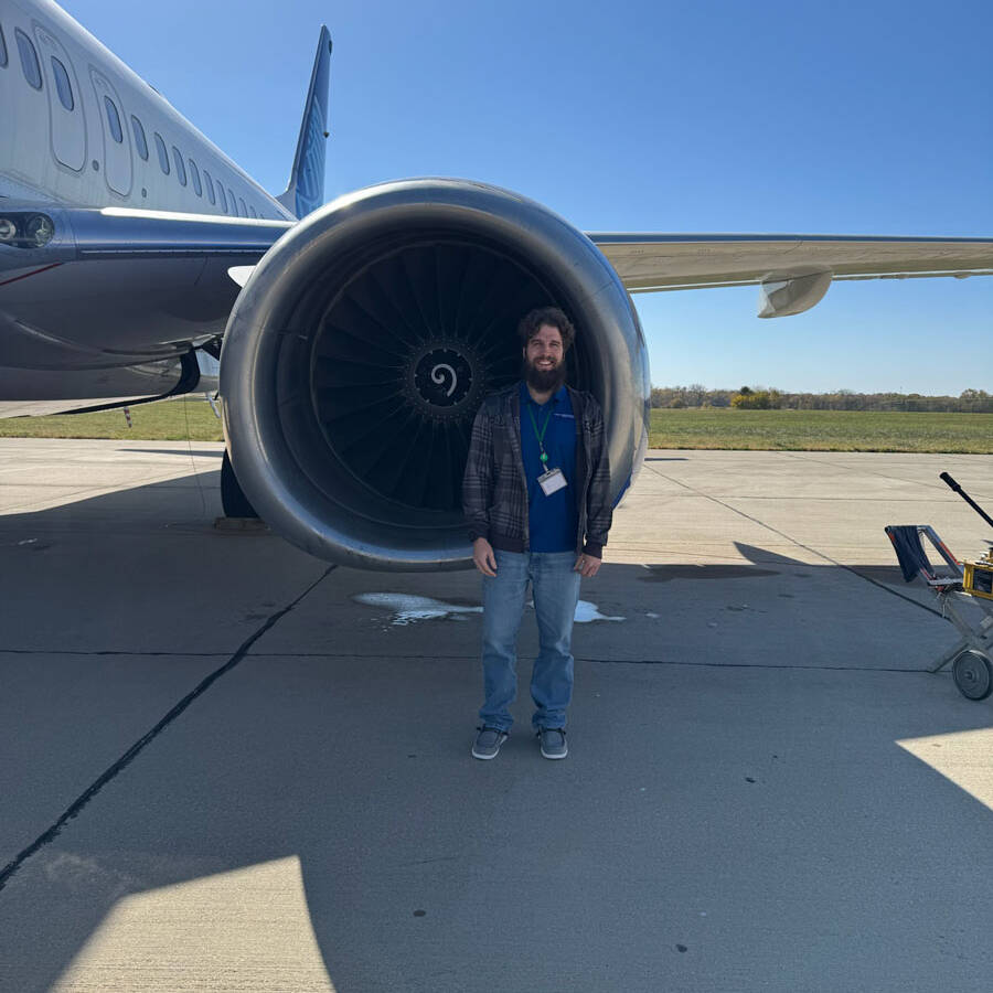 A person stands in front of a large airplane engine on the sunny airport tarmac, wearing casual clothing and a lanyard. The clear sky reflects the excitement of air travel. Part of the airplane's body and wing loom nearby, ready for their next visit to explore destinations like Rockport.