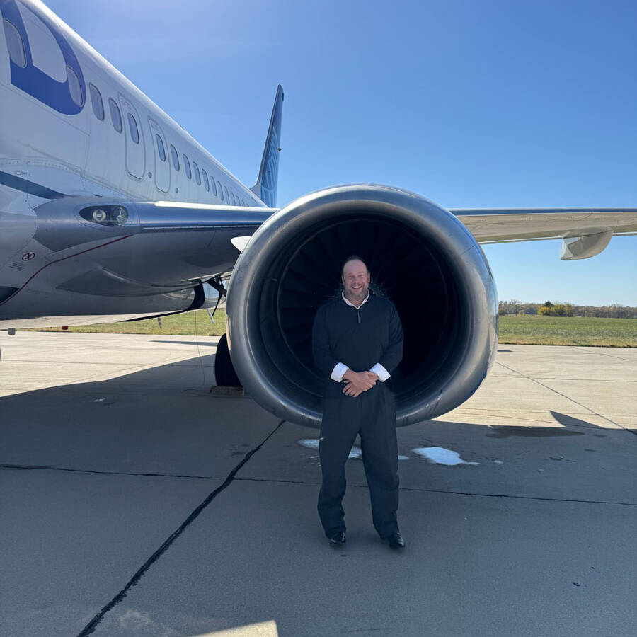 A man stands smiling in front of a large jet engine on a sunny day at Rockport Airport, with the airplane parked on the tarmac behind him. Wearing a dark sweater and pants, he enjoys his visit under the clear blue sky.