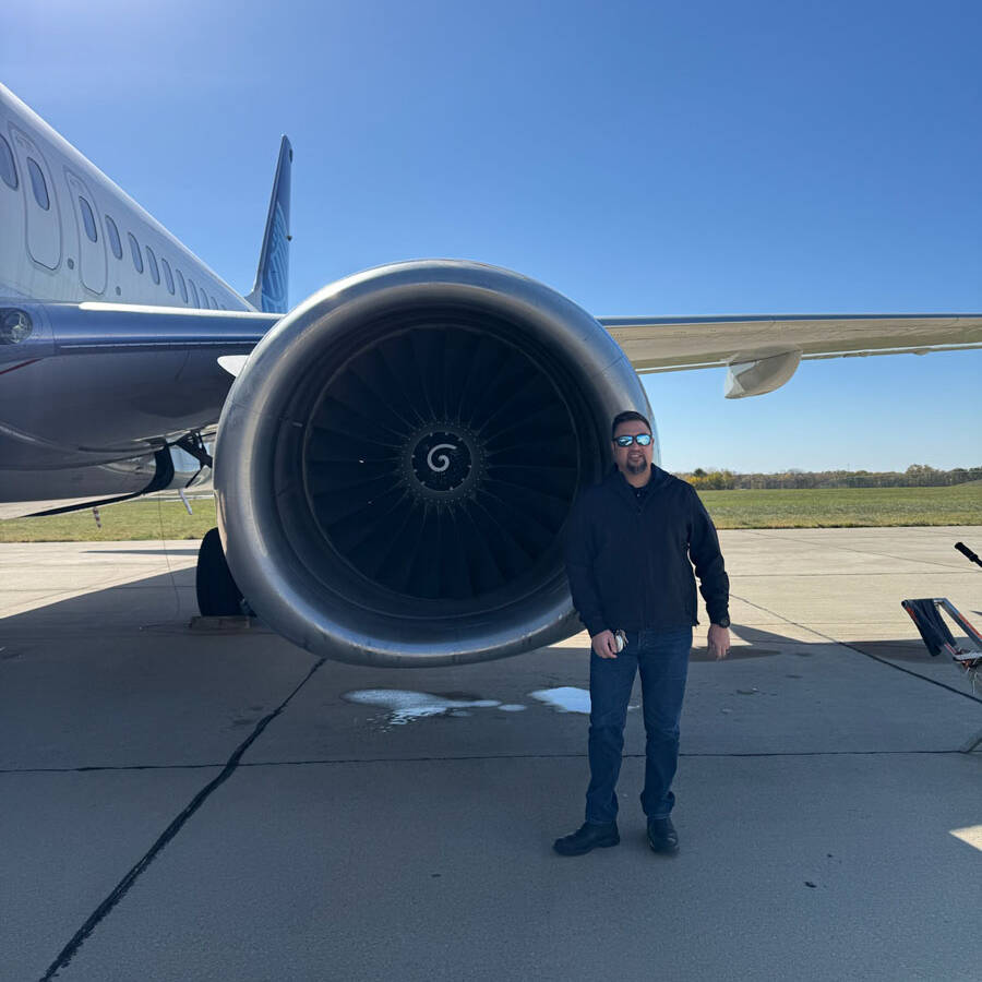 A person stands on an airport tarmac in front of a large jet engine, poised for their next adventure. Sporting sunglasses, a black jacket, and jeans, they embody the spirit of travel. A vibrant blue sky frames the scene alongside the aircraft's tail section as they prepare to visit Rockport.