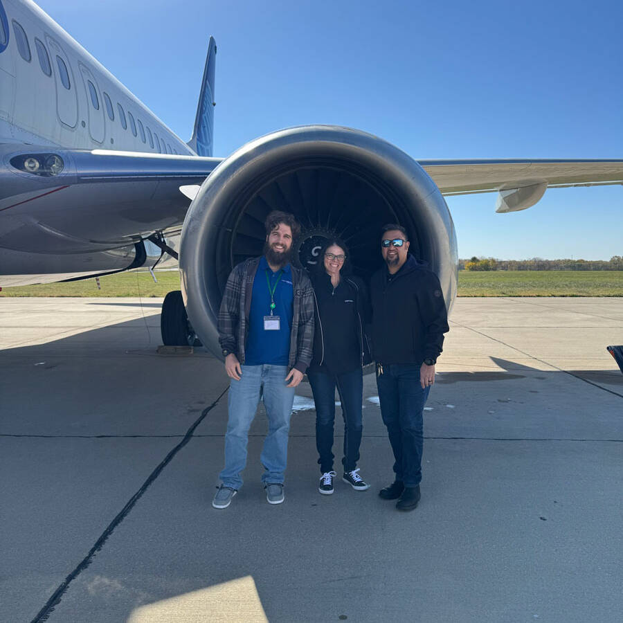 Three people stand in front of a large airplane engine at Rockport Airport, with part of the aircraft visible on the left. It's a sunny day with a clear blue sky, making it the perfect visit spot for aviation enthusiasts.