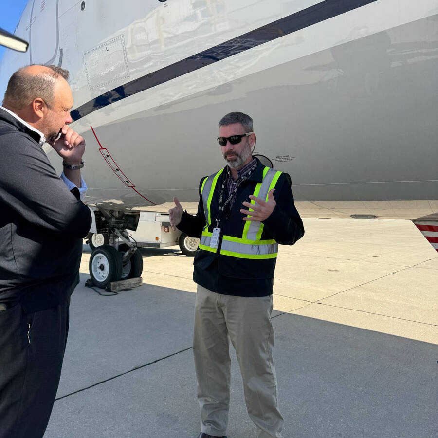 A man in a neon safety vest stands on an airport tarmac beside an airplane, gesturing with his hands. Another man in a black jacket listens with his hand on his chin, planning their visit. The airplane's fuselage looms in the background.