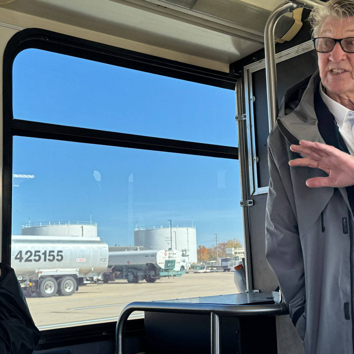 An older individual speaking and gesturing while standing inside a bus near Rockport. Another person is seated nearby, listening intently. Outside the window, a tanker truck with the number 425155 is parked near large storage tanks under the clear blue sky.