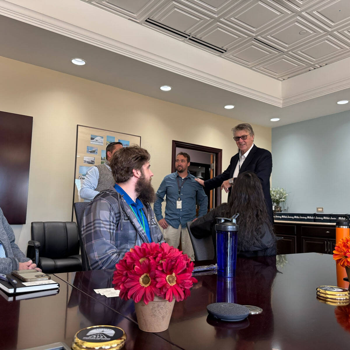 A group of people gathered around a conference table in an office near the bustling airport. One person stands, speaking to the others. The table is decorated with red and orange flowers, and a wall-mounted logo is visible in the background.