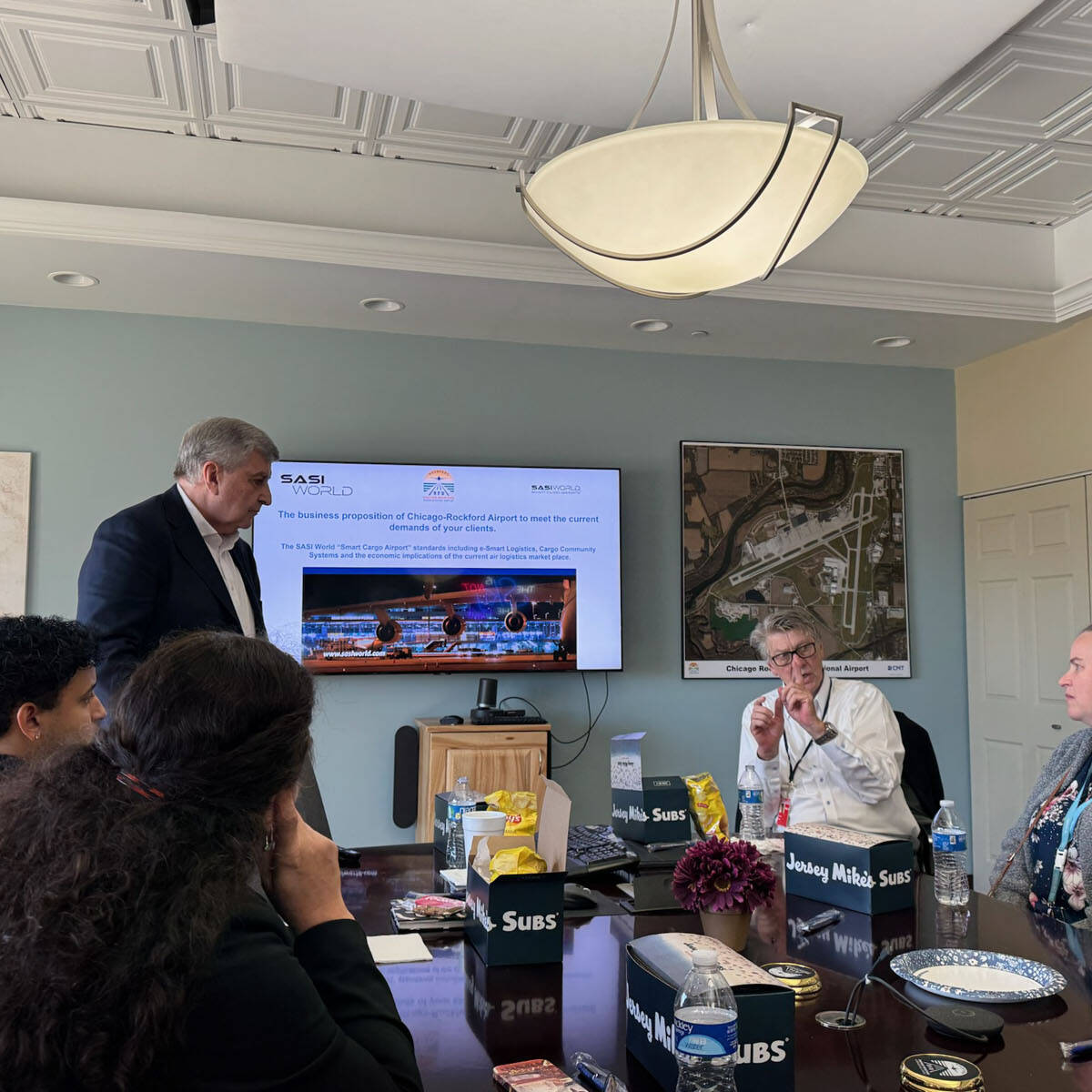 A group of people are sitting around a conference table in a meeting room. A man is standing and giving a presentation while a screen displays information. Various items, including food containers from Jersey Mike's Subs, are on the table, adding local flavor to this pre-visit itinerary discussion for Rockport.