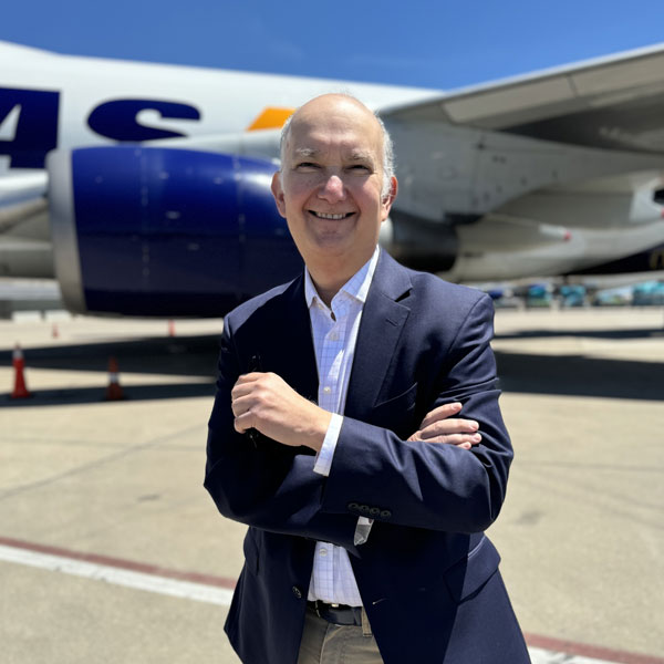 A person standing confidently with arms crossed in front of an airplane on a sunny day. The airplane has a large jet engine visible. The person is wearing a dark blazer and light shirt, smiling with a clear blue sky in the background.