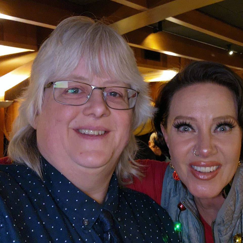 Two people smiling for a selfie at the 2024 Holiday Party in a warmly lit indoor setting. The person on the left has gray hair and glasses, wearing a dark dotted shirt, while the person on the right sports short dark hair and a colorful vest with ornaments. A bar area is in the background.