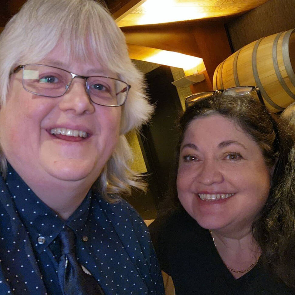 Two people smiling indoors at a holiday party, with wooden barrels in the background. One person has light-colored hair and glasses, wearing a polka dot shirt. The other has dark hair and is wearing a dark top. The setting appears warm and cozy, embracing the festive spirit of 2024.