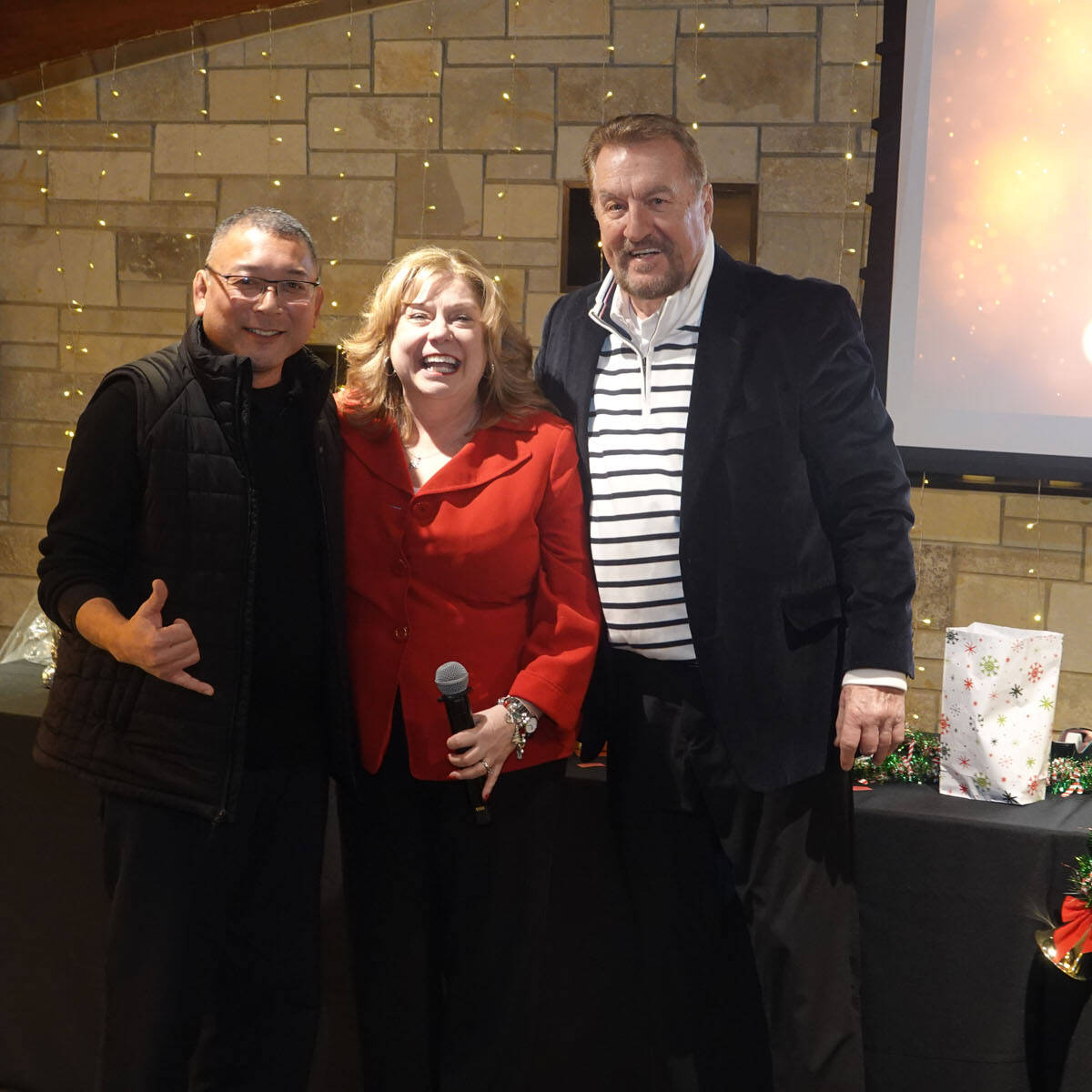Three people stand together, smiling in front of a stone wall decorated with string lights, exuding party vibes. The person on the left gestures with a hand sign, the one in the middle holds a microphone as if announcing "Happy Holidays 2024," and the person on the right rests an arm on their shoulder.