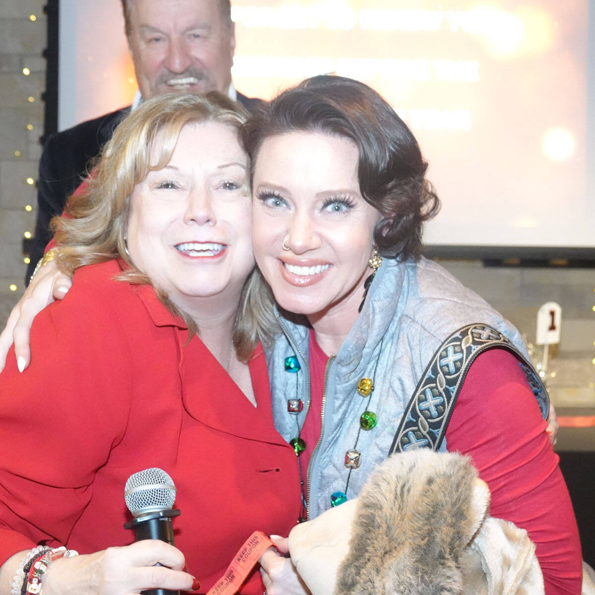 Two women, one holding a microphone, pose happily together at a 2024 holiday party. They stand in front of a wall with a projected screen, while a man smiles behind them. Festive lights and colorful gift bags add to the vibrant atmosphere.