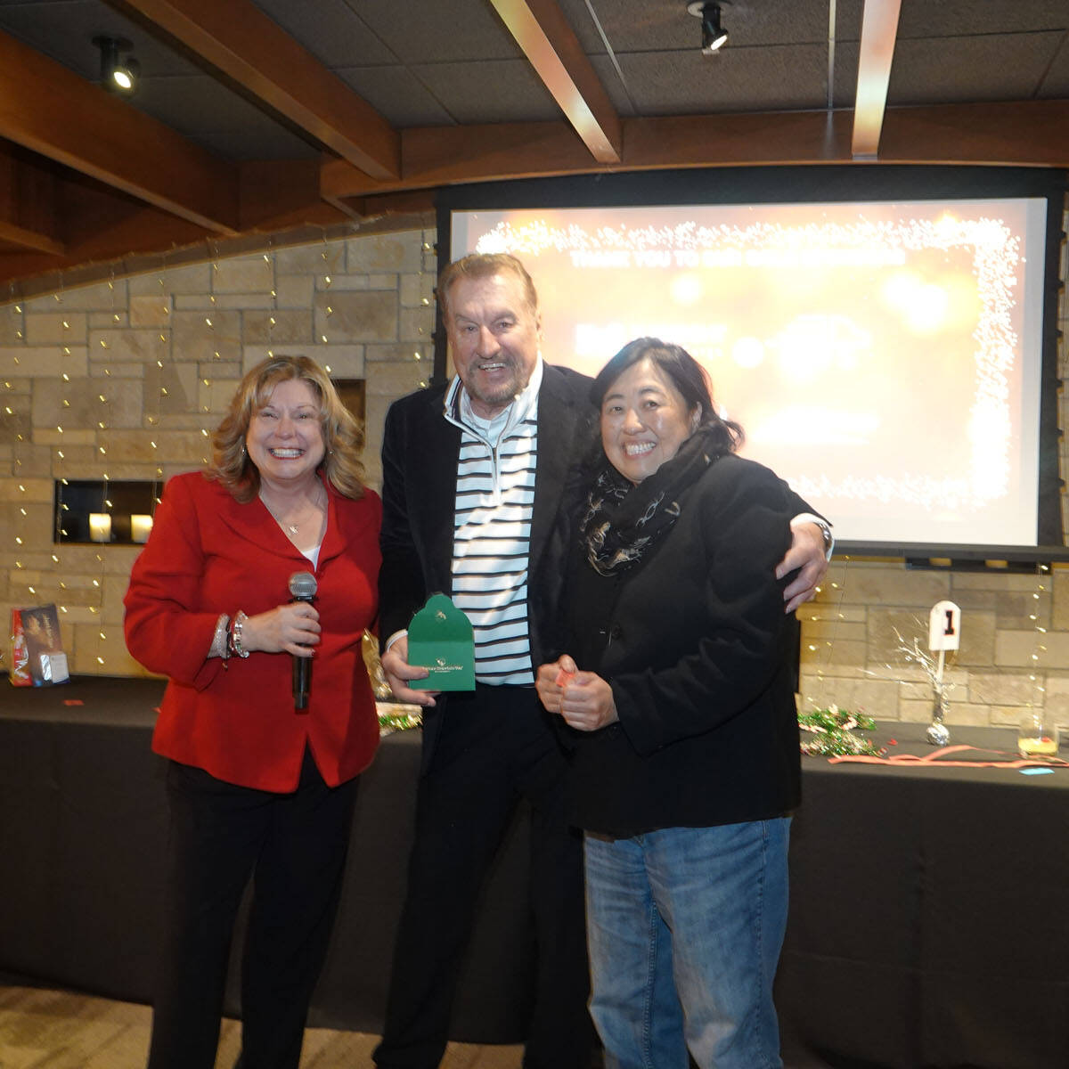 Three people stand together in a festive setting, likely at a holiday party. The person in the middle holds a green award marked with "2024," smiling against the backdrop of a decorated table and a large screen.