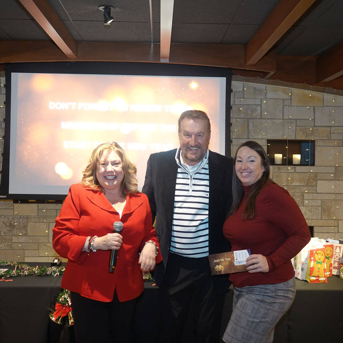 Three people are standing indoors, smiling at the camera during a holiday party. The woman on the left holds a microphone. Behind them is a screen displaying an unreadable message and a wall with candles, adding to the festive vibe of this 2024 event, with holiday-themed decorations visible on a table.