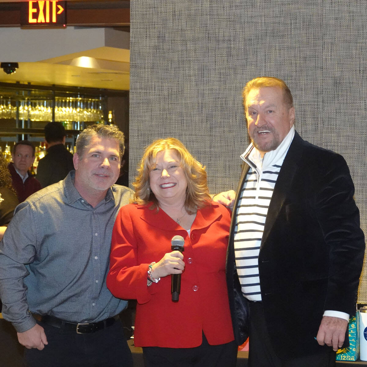 Three people stand together in a lively restaurant setting, smiling for the camera at a festive holiday party. The woman in the center holds a microphone, wearing a red blazer. The men beside her wear casual and semi-formal attire, with others mingling in the 2024 celebration background.
