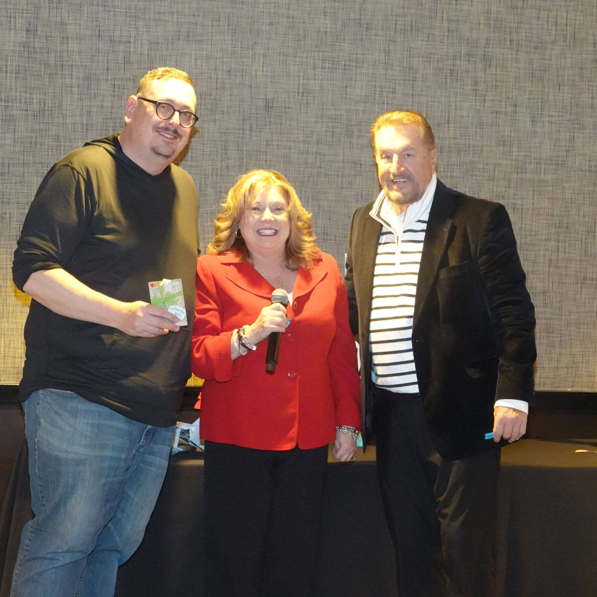 Three people are standing together indoors at a festive party. The person in the center, holding a microphone and wearing a red jacket, exudes holiday spirit. The person on the left holds a small item, while the one on the right is in a suit. All are smiling warmly towards the camera, embracing 2024 vibes.