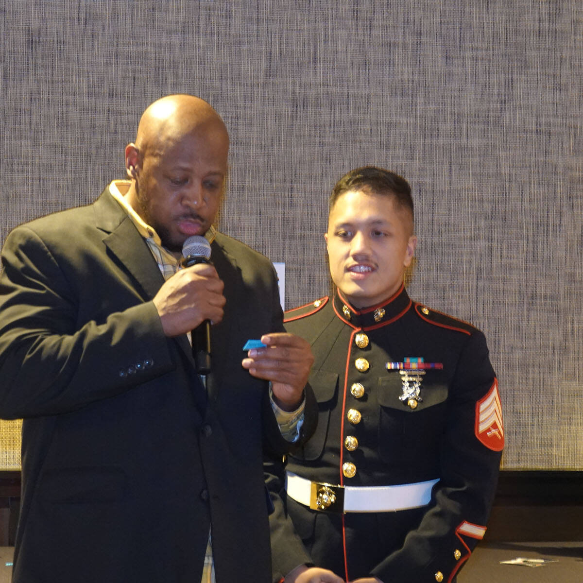 Two men are standing indoors at a formal event. The man on the left, in a black suit, speaks into a microphone while holding a small blue item. The man on the right, dressed in a military uniform adorned with medals, observes attentively. It's clear this gathering is part of an early 2024 celebration.